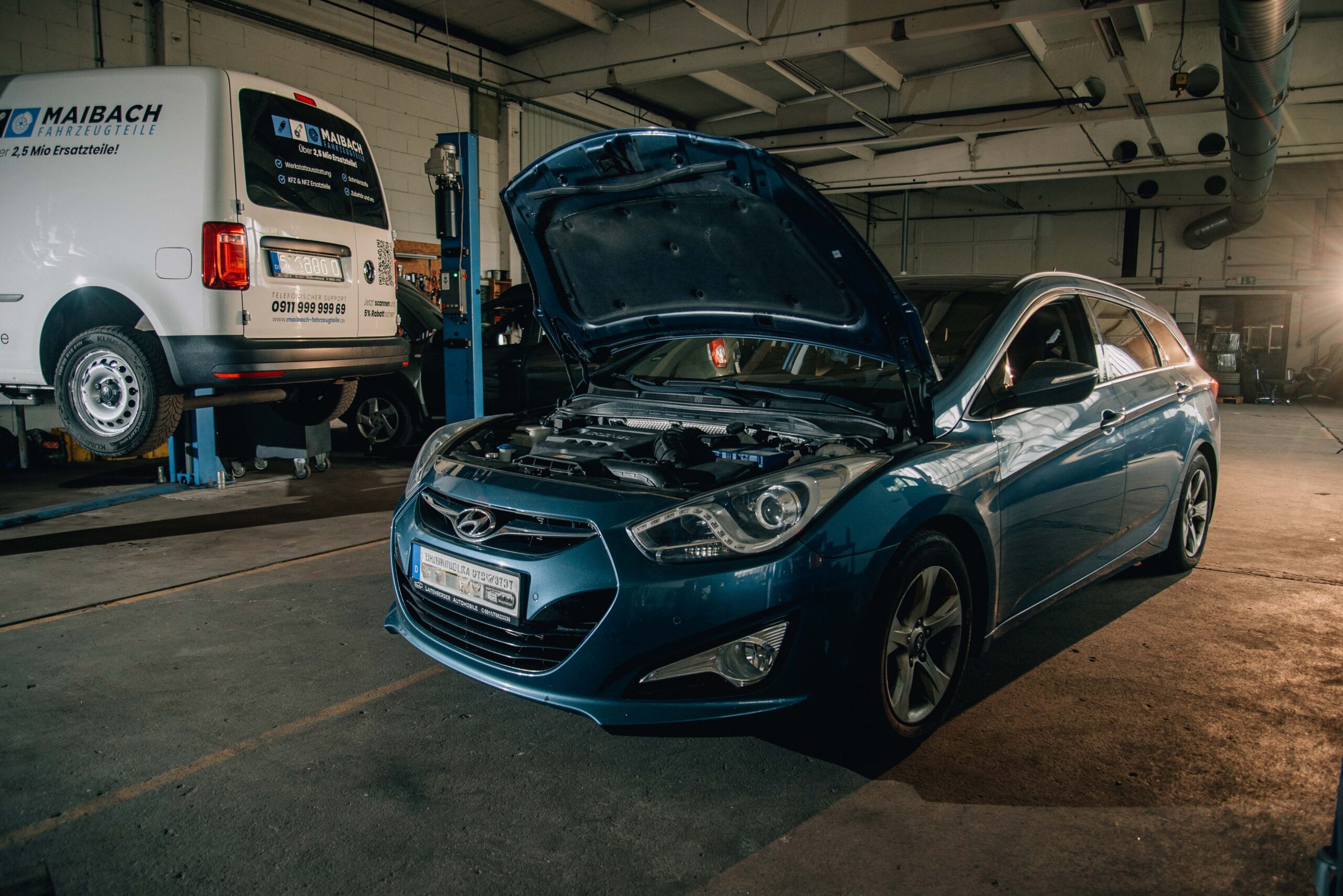 Ein blauer Hyundai mit geöffneter Motorhaube steht in einer Autowerkstatt. Im Hintergrund ist ein weißer Lieferwagen mit der Aufschrift "MAIBACH Fahrzeugteile" auf einer Hebebühne zu sehen. Die Werkstatt ist gut beleuchtet und zeigt verschiedene Werkzeuge und Ausrüstungen